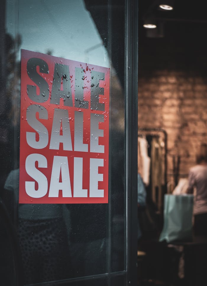 About Red sale sign on a glass door with interior view of a clothing store.